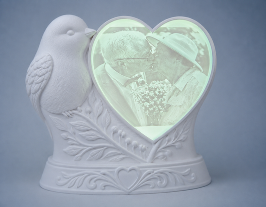 A white decorative ceramic figurine shaped like a bird perched on a leafy branch, with a heart-shaped photo window showing a couple kissing. The base is embossed with floral and heart motifs, set against a soft blue-gray background.